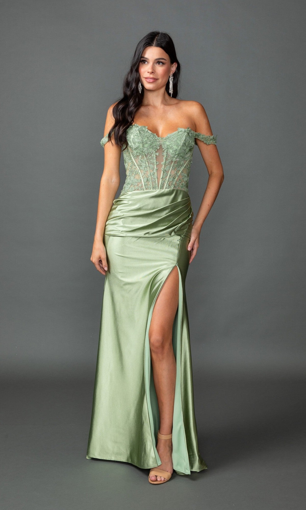 Wearing the Dancing Queen 4536, a light green, off-shoulder, floor-length prom dress with lace bodice, sheer paneling, and thigh-high slit, a woman poses against a plain gray background.